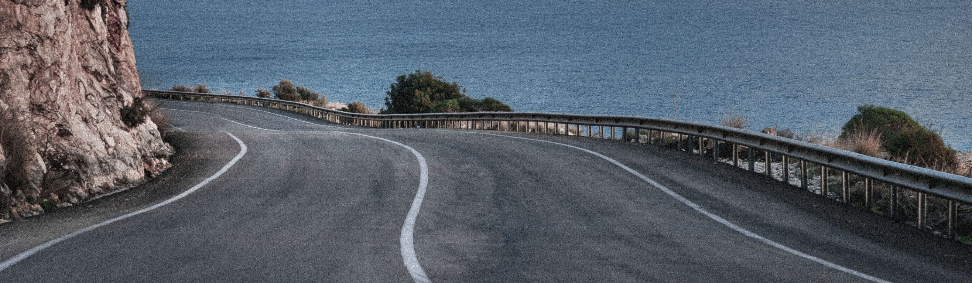 Image from MUDDY VIBE. A winding road with guardrails beside it, bordered by a rocky cliff and the sea.