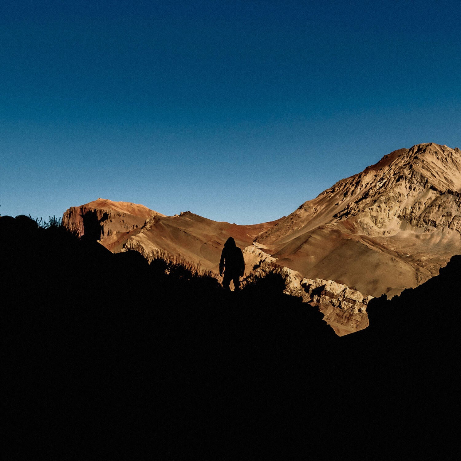 Image from MUDDY VIBE. A person stands silhouetted against a backdrop of majestic mountains under a clear sky.