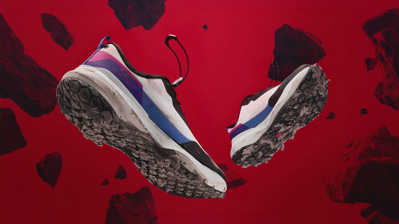 MUDDY VIBE Si-14 low-top Aurora White hiking shoe floating mid-air with dirt on sole, rocks gently moving on red background, side view.