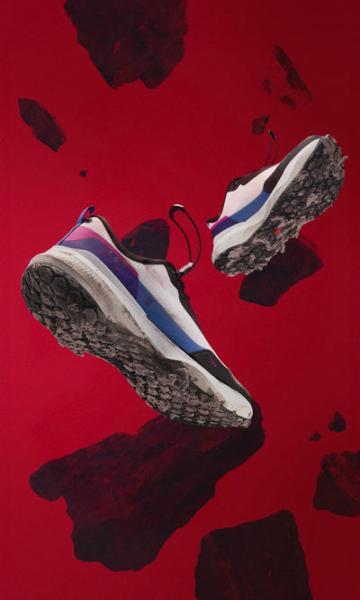 MUDDY VIBE Si-14 low-top Aurora White hiking shoe floating mid-air with dirt on sole, rocks gently moving on red background, side view.
